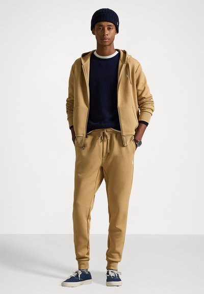 Tan zip-up hoodie with front pockets, paired with a navy sweatshirt and matching joggers. Accessories include a ribbed beanie and navy sneakers.