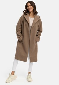 Manteau long brun clair avec capuche, en tissu doux, offrant une coupe décontractée et des poches latérales. Associé à un pantalon blanc et des baskets légères.