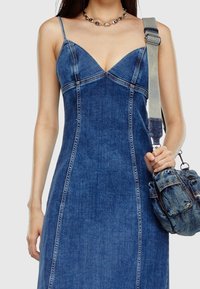 Kvinna iklädd en åtsittande blå denimklänning med tunna band, håller en matchande denimaxelväska med en justerbar rem.