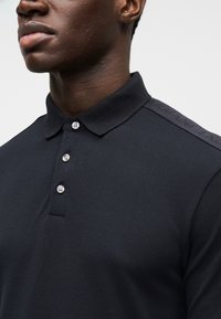 Tumesinine polo särk tekstuurse kangaga, millel on kolmenupu plastron ja õlal reljeefne logo. Klasikalise krae disain.