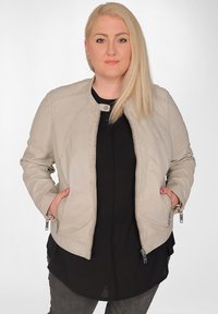 Beige bomberjacka med dragkedja och rund hals, med sidofickor och justerbara dragkedjor på ärmarna, bärs över en svart topp.