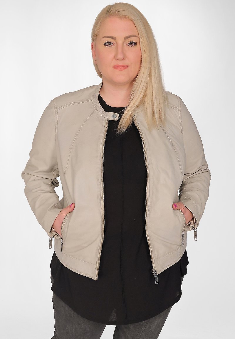 Beige bomberjacka med dragkedja och rund hals, med sidofickor och justerbara dragkedjor på ärmarna, bärs över en svart topp.
