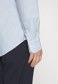 Camicia azzurra chiara con orlo curvo e polsini abbottonati, abbinata a pantaloni scuri in navy con texture. Tessuto morbido con un leggero lucido.