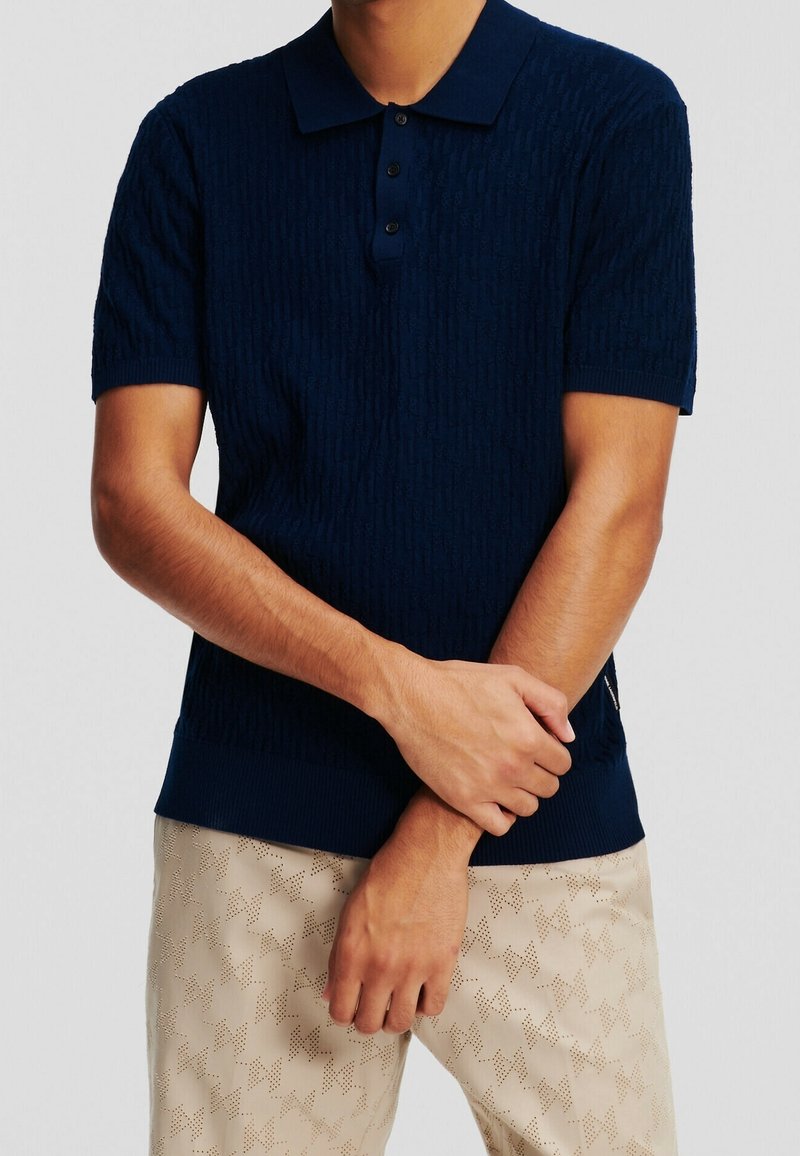 Homme portant un polo en maille bleu marine à manches courtes et un pantalon beige avec un motif géométrique subtil, les bras croisés devant.