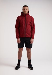 Rode waterdichte jas met een voorrits, verstelbare capuchon en twee zijzakken. Draag met zwarte korte broek en zwarte sport schoenen.