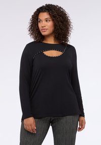 Žena s kudrnatými vlasy, oblečená v černém triku s dlouhým rukávem, které má výstřih s hřebíčky nad hrudníkem, a s texturovanými šedými kalhotami.