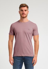 Kurzärmeliges T-Shirt mit Rundhalsausschnitt aus schlichten Mauve Baumwolle. Enthält ein kleines, quadratisches Logo auf der Brust. Getragen mit blauen Denim-Jeans.