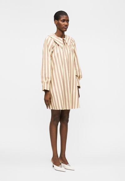 Γυναίκα που φοράει φόρεμα με κάθετες ρίγες σε μπεζ και κρεμ αποχρώσεις, μακριά μανίκια και μεγάλο γιακά, συνδυασμένο με λευκές μυτερές γόβες.