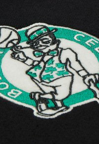 Parche bordado que representa un duende de los duendes en un diseño verde y blanco, con una pajarita, sosteniendo un balón de baloncesto sobre un fondo negro.
