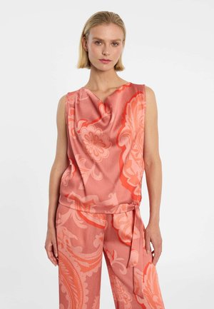 Vrouw draagt een mouwloze koraalroze top en bijpassende wijde broek met grote bloemenpatronen, staand tegen een effen witte achtergrond.