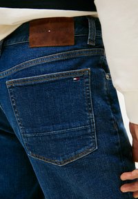 Jeans in denim blu scuro con un'etichetta in pelle sul retro con il marchio "TOMMY HILFIGER", cuciture visibili e un piccolo logo sulla tasca posteriore.