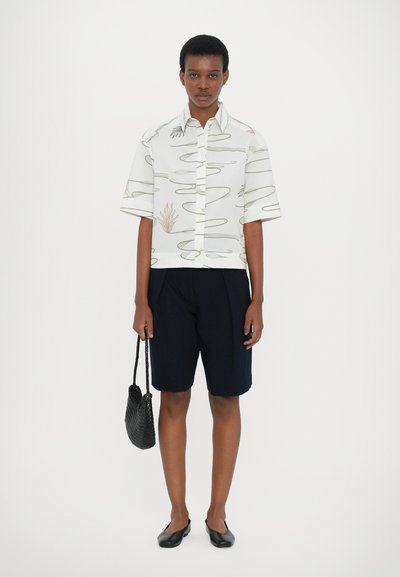 Short-sleeve white shirt with abstract green and beige patterns, paired with navy shorts; black woven bag and flat shoes complete the look.