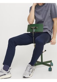 Grön metallrullstol med en böjd ryggstöd, marinblå byxor, grå t-shirt och grå sneakers. Släta texturer är synliga.