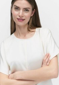 Biała bluzka z krótkim rękawem, wykonana z gładkiego materiału, z okrągłym dekoltem i szwami z przodu. Modelka pozuje z założonymi rękoma.