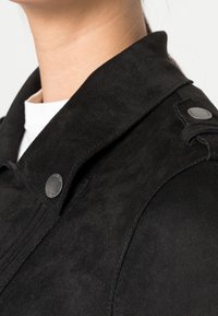 Veste en daim noire avec un large col, des boutons-pression sur l'épaule, et une texture lisse. Vue rapprochée montrant les détails du tissu et les coutures.