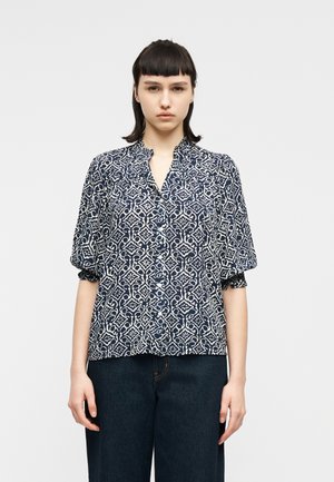 Młoda osoba z krótkimi ciemnymi włosami, ubrana w granatowo-białą koszulę zapinaną na guziki z kwiatowym wzorem oraz ciemne jeansy, stojąca na jednolitym tle.