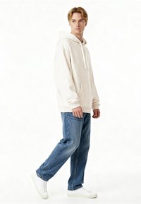 Sudadera con capucha de color beige claro, hecha de material suave, con un bolsillo canguro y puños de canalé, combinada con jeans de denim azul sueltos y zapatillas blancas.