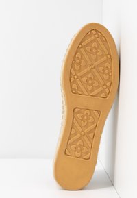 Semelle d'une chaussure taille 37 couleur caramel avec des motifs floraux et géométriques sur la bande de roulement, reposant contre un mur d'angle blanc sur une surface blanche.
