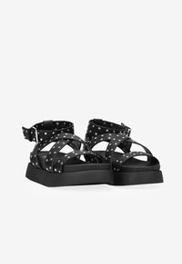 Black sandals with cross straps, metallic stud accents, and a chunky black sole. Adjustable buckle at the ankle for secure fit.