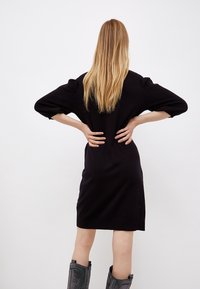 Abito nero in maglia con una vestibilità rilassata, maniche a sbuffo tre quarti e orlo a coste. Modella che indossa stivali grigi testurizzati fino al ginocchio.