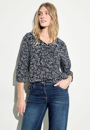 Blouse marine avec un motif paisley blanc, manches trois-quarts et détail à lacets. Assortie à un jean en denim foncé. Tissu structuré et coupe décontractée.