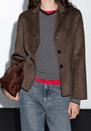 Blazer en laine marron à trois boutons, assorti à une chemise rayée noir et blanc et un jean bleu clair. Tenant un petit sac en cuir marron.