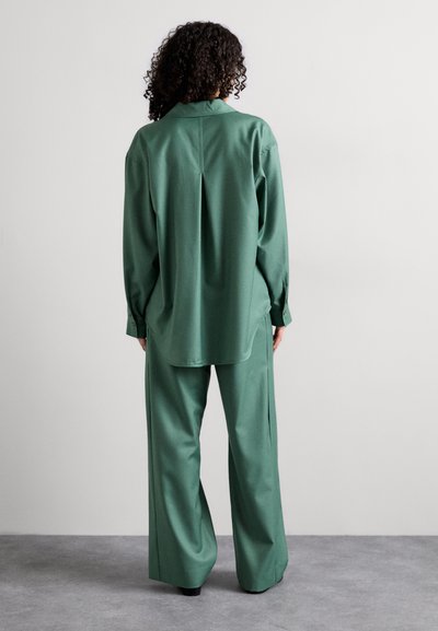 Camicia verde, ampia e comoda, con colletto e piega sul retro, abbinata a pantaloni larghi realizzati nello stesso materiale e colore. Tessuto liscio.