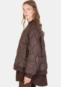 Giovane donna con capelli lunghi che indossa una giacca trapuntata marrone scuro larga e un completo a strati marrone abbinato, in piedi di profilo.