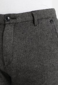 Pantalons à motif chevrons gris avec une taille plate, featuring un fermoir à bouton noir et des passants de ceinture, fabriqués en tissu texturé.