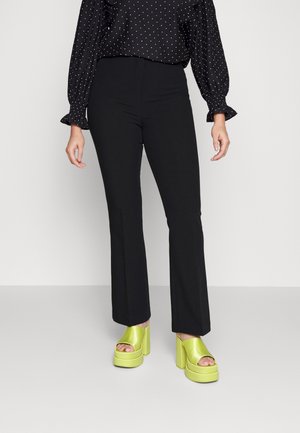 Femme portant un pantalon noir ajusté, un chemisier noir à pois à manches longues, et des sandales à talons hauts plateforme vert citron, debout sur un sol gris.
