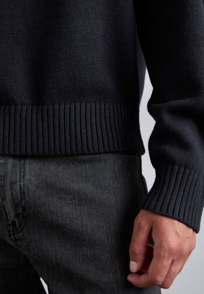 Navy knit sweater with ribbed cuffs and hem, paired with textured gray jeans. The sweater features a smooth finish and relaxed fit.