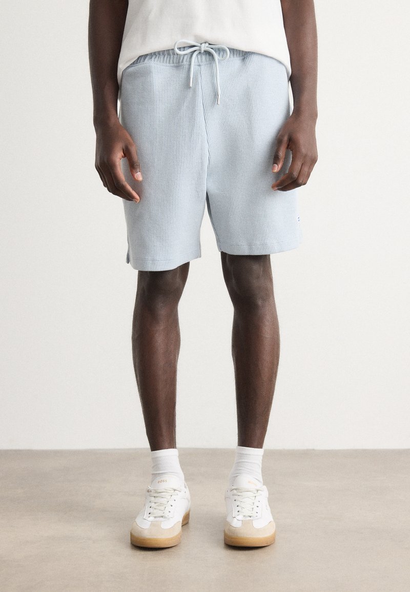 Light blue ribbed shorts with elastic waistband and drawstring. Features side vents and logo accent. Worn with white sneakers and socks.