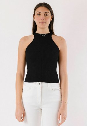 Donna con capelli lunghi e scuri che indossa un top nero senza maniche a coste con scollo all'americana e pantaloni bianchi a vita alta, in piedi davanti a uno sfondo semplice.