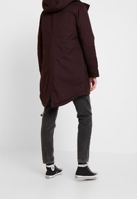 Cappotto lungo color bordeaux scuro con cappuccio, caratterizzato da un taglio dritto, due tasche laterali e un orlo più lungo sul retro. Indossato con jeans grigi e scarpe da ginnastica nere.