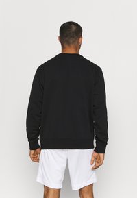 Uomo con capelli corti che indossa una maglietta nera a maniche lunghe e pantaloni bianchi, in piedi con la schiena rivolta verso la macchina fotografica su uno sfondo semplice.