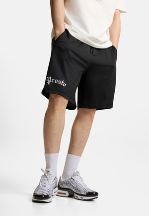 Osoba ubrana w czarne spodenki z napisem „Prosto” na lewej nodze, białe skarpetki i białe oraz czarne wzorzyste sneakersy, stojąca z rękami w kieszeniach.