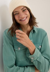 Femme souriante portant un bonnet en tricot crème et une veste bleu canard à boutons, avec une main touchant son col devant un fond clair et uni.