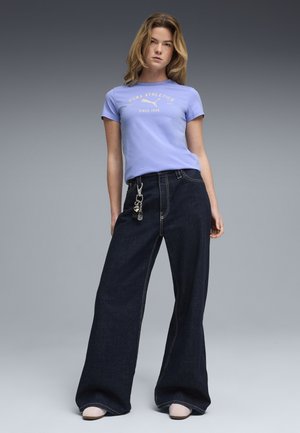 Donna in piedi che indossa una maglietta da atletica Puma color lavanda, jeans scuri a gamba larga con portachiavi e scarpe chiare su sfondo grigio.