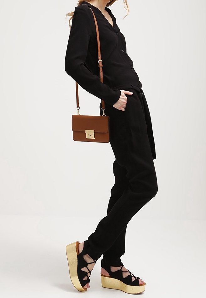 Femme portant un haut noir boutonné et un pantalon, portant un sac à épaule marron, portant des sandales compensées à lacets noires avec semelles en liège.