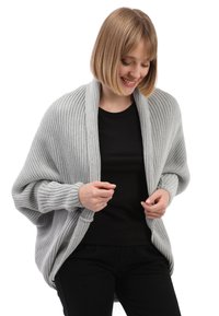 Lichtgrijze ribgebreide cardigan met een gedrapeerd ontwerp. Heeft een sjaalkraag en aansluitende mouwmanchetten, gedragen over een zwarte top.