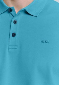 Turquoise poloshirt gemaakt van gestructureerde stof, met een drieknopenlijst en een subtiele NXS-logo op de borst.