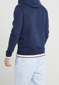 Felpa blu navy con un design attillato, caratterizzata da un grande cappuccio e polsini e orlo a coste con righe bianche. Indossata con pantaloni azzurri.