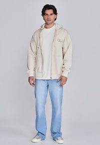 Beige Sweatshirt mit Reißverschluss und weißem Innenfutter, kombiniert mit weißem T-Shirt und hellblauen Jeans mit geradem Bein. Weiße Sneaker vervollständigen das Outfit.