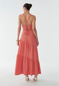 Maxi sukienka w kolorze koralowym, z teksturowanej tkaniny. Posiada wzór ze sznurówką z tyłu oraz falbaniastą spódnicę. Ramiączkowy krój z odkrytymi plecami. Elegancka sylwetka.