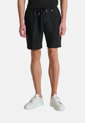 Muškarac u crnim kratkim hlačama na vezanje i bijelim tenisicama stoji ispred jednobojne svijetle pozadine.