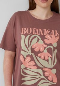 T-shirt marron avec un motif botanique présentant des fleurs roses et des feuilles vertes. Col rond et manches courtes. Texture de tissu douce.