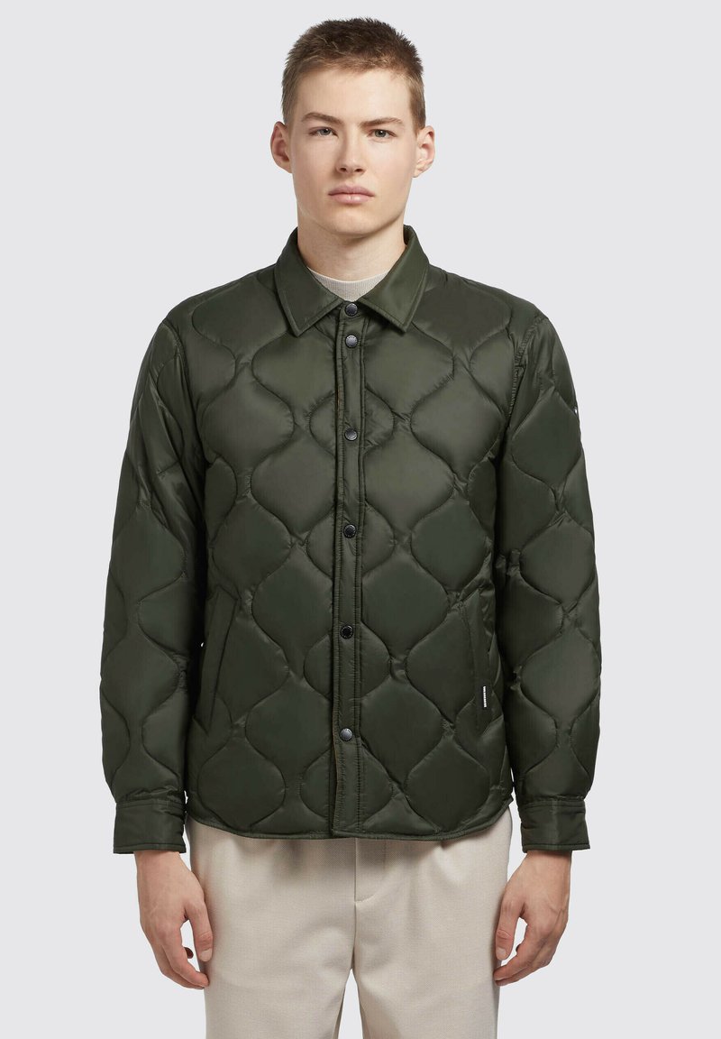 Gevulde groene jack met een gequiltte textuur, met drukknopen aan de voorkant en een kraag. Draagt over een lichtgekleurde shirt.