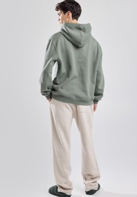 Sweat à capuche en tissu vert doux, coupe décontractée. Associé à un pantalon de jogging beige clair, montrant des poches latérales et un design décontracté.