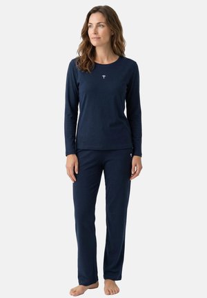 Kvinde står barfodet iført en navyblå langærmet top og matchende bukser med små hvide logoer på bryst og lår.