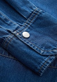 Denim shirt cuff featuring a silver button, mustard stitching, and a smooth texture, with a deep blue color and folded design.